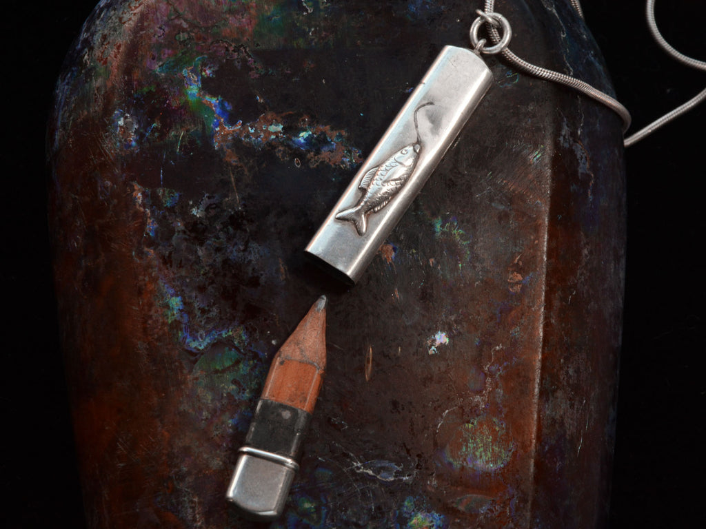 Detail of silver pendant with fish decoration, shown open with concealed wooden pencil inside, on dark background.