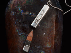 thumbnail of Detail of silver pendant with fish decoration, shown open with concealed wooden pencil inside, on dark background.