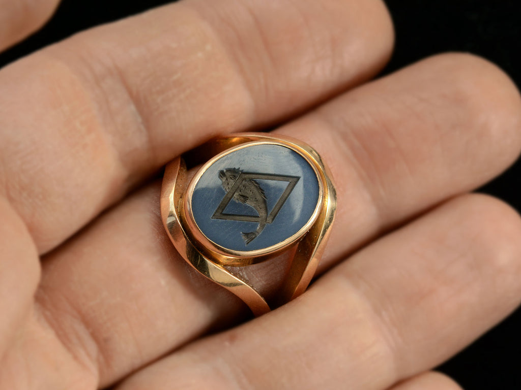 A vintage French yellow gold signet ring with the image of a fish jumping through a lozenge shape carved into blue and black chalcedony. Shown on hand.