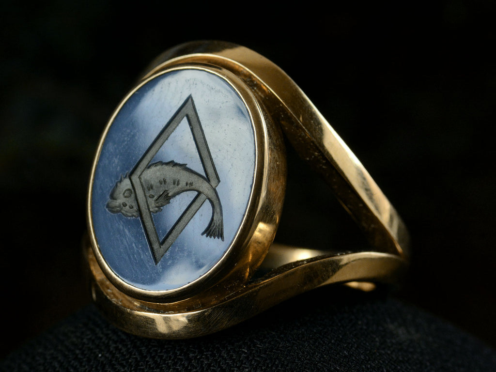 Right angled front view of a vintage French yellow gold signet ring with the image of a fish jumping through a lozenge shape carved into blue and black chalcedony. Shown on dark background.