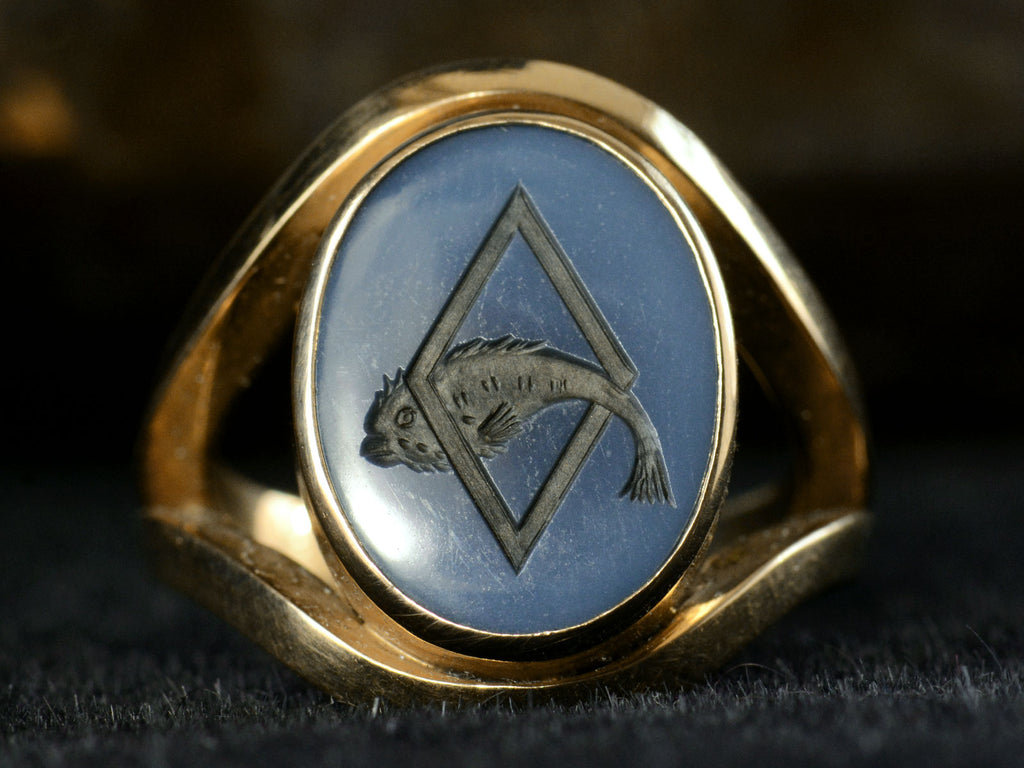 Front view of a vintage French yellow gold signet ring with the image of a fish jumping through a lozenge shape carved into blue and black chalcedony. Shown on dark background.