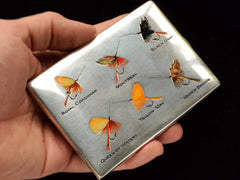 thumbnail of Hand holding silver cigarette case with fly fishing lures labeled 'Royal Coachman', 'Montreal', 'Black Gnat', 'Queen of Waters', 'Yellow May', and 'March Brown' on a black background.