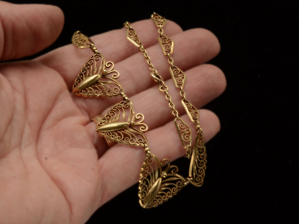 An antique gold filigree chain necklace with three large decorative leaf-like pendants, held in a person's hand against a black background.