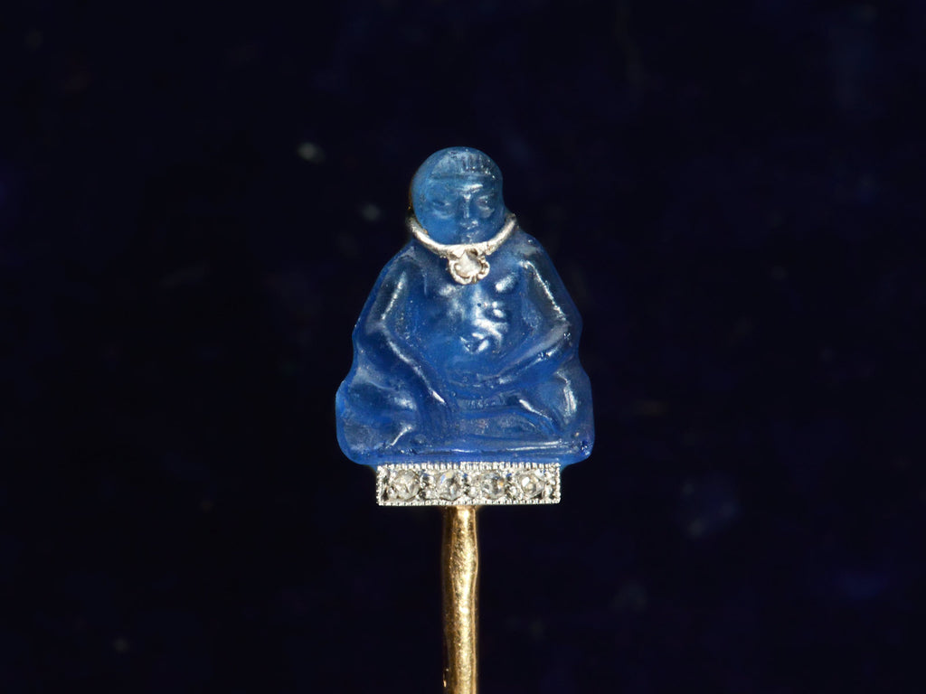 Detail of French stick pin with blue glass Buddha decorated with diamonds in platinum and yellow gold, shown on dark background.