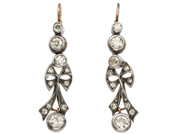 A pair of c1890 Victorian drop earrings with old European cut diamonds set in silver topped gold with a bow design. Shown on white background.