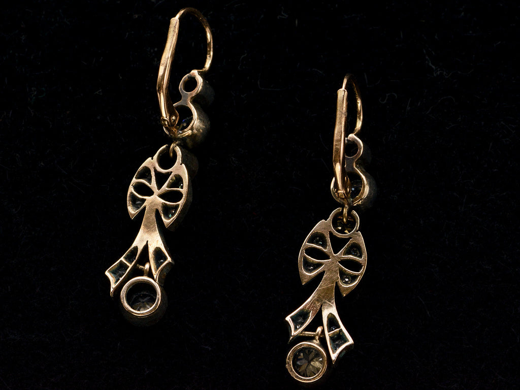 Reverse sides of a pair of c1890 Victorian drop earrings with old European cut diamonds set in silver topped gold with a bow design. Shown on black background.