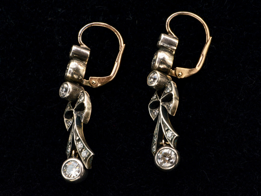Side view of a pair of c1890 Victorian drop earrings with old European cut diamonds set in silver topped gold with a bow design. Shown on black background.