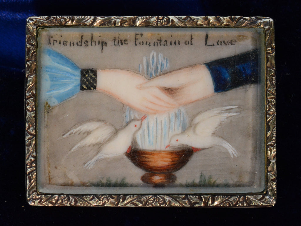 An antique Georgian-era rectangular brooch with hands and doves and the words "Friendship the Fountain of Love" with a gold frame. Shown on dark blue background.