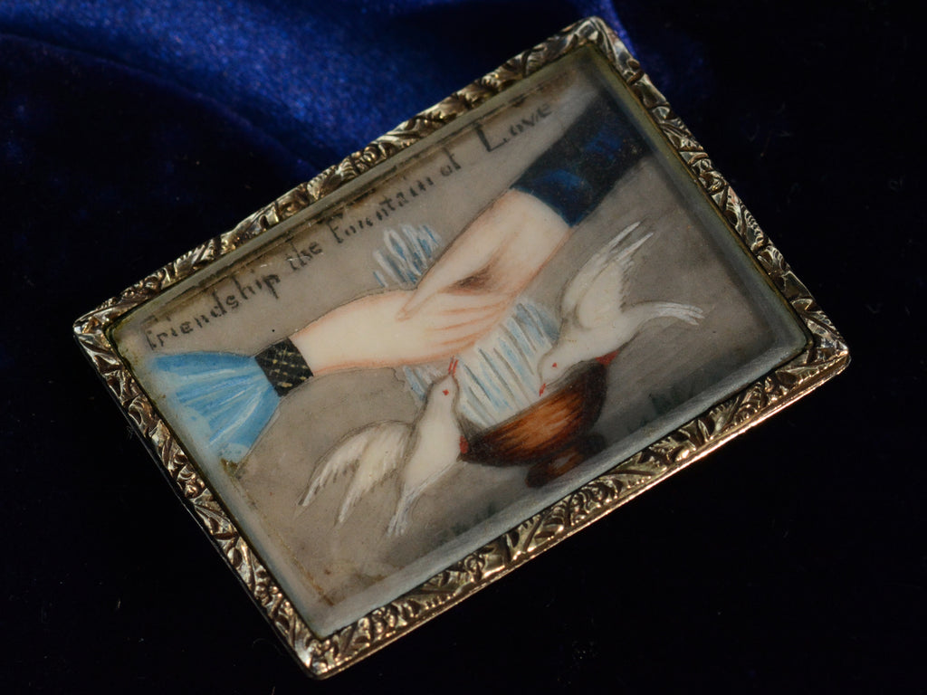 Angled view of an antique Georgian-era rectangular brooch with hands and doves and the words "Friendship the Fountain of Love" with a gold frame. Shown on dark blue background.