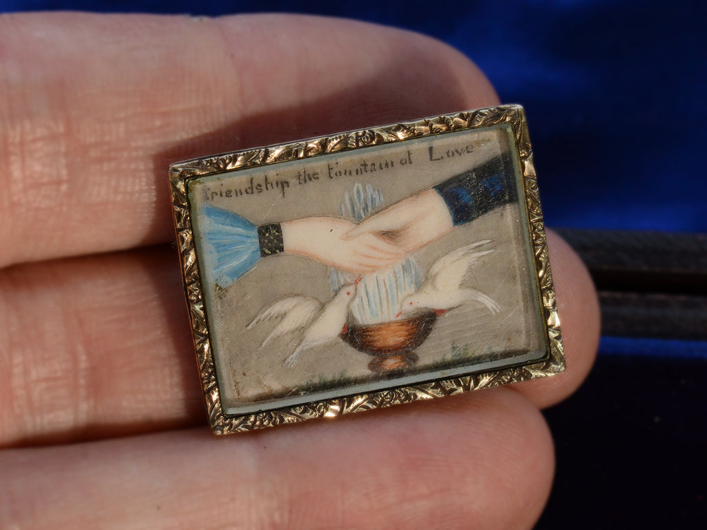 An antique Georgian-era rectangular brooch with hands and doves and the words "Friendship the Fountain of Love" with a gold frame. Shown on hand for scale.