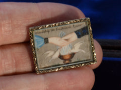 thumbnail of An antique Georgian-era rectangular brooch with hands and doves and the words "Friendship the Fountain of Love" with a gold frame. Shown on hand for scale.