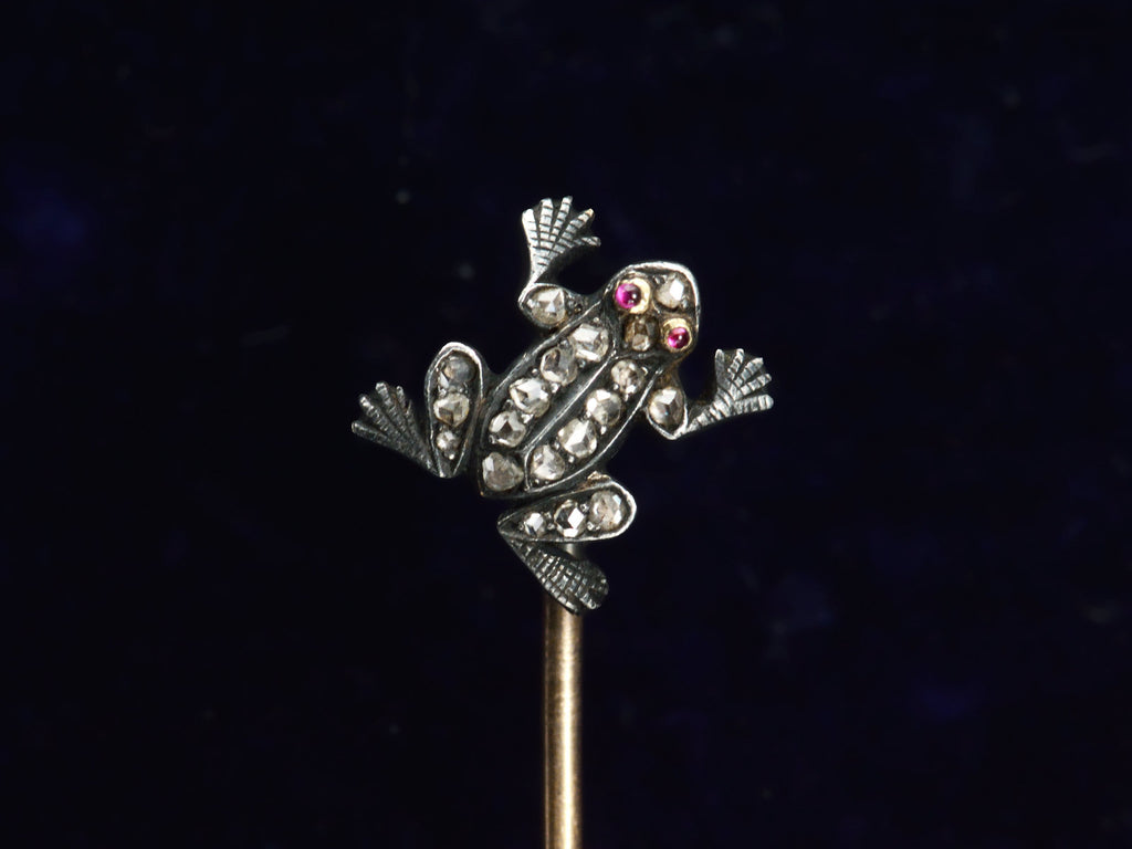 Detail of antique frog stick pin in silver and gold with rose cut diamonds and ruby eyes, shown on dark background.