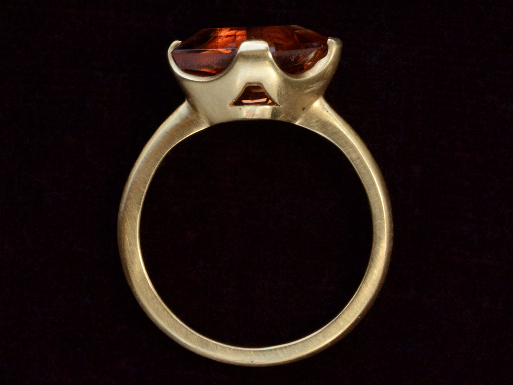 Side profile view of shield shaped orangey red stone set in 18K yellow gold, on dark background.