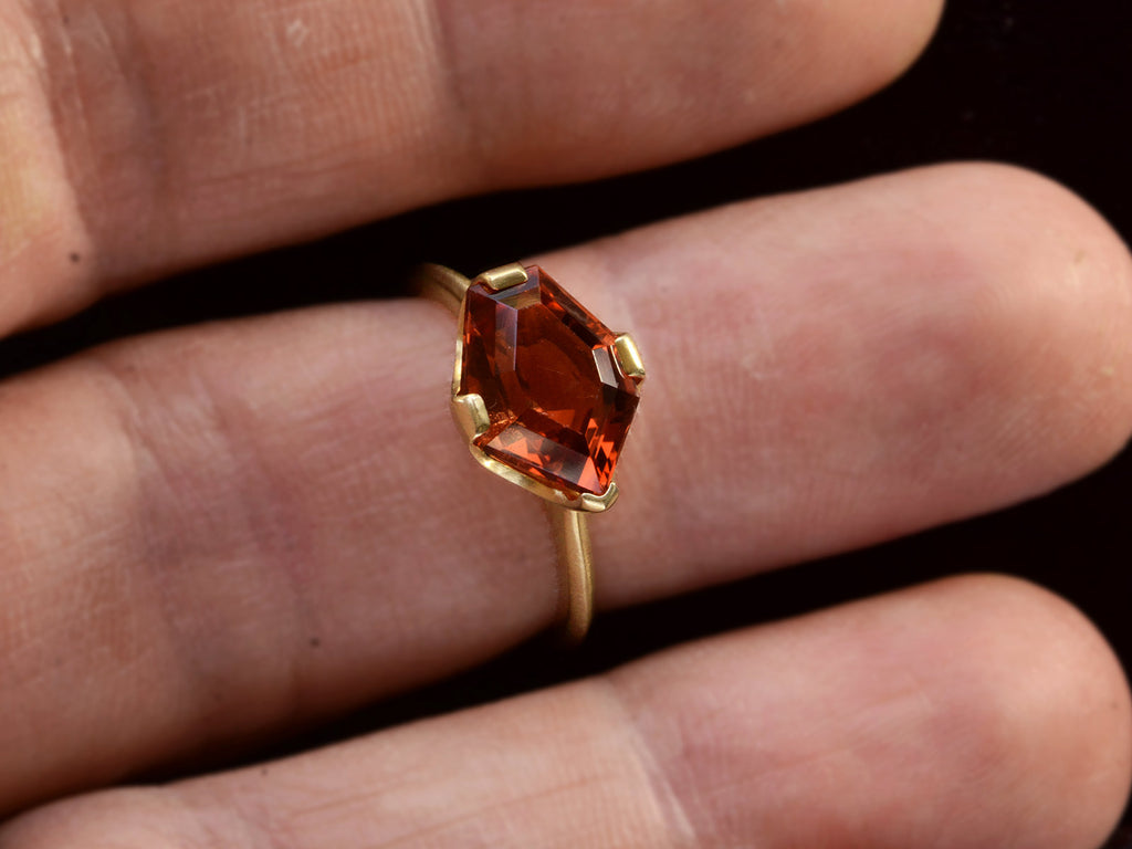 Face on view of shield shaped orangey red stone set in 18K yellow gold, on finger.