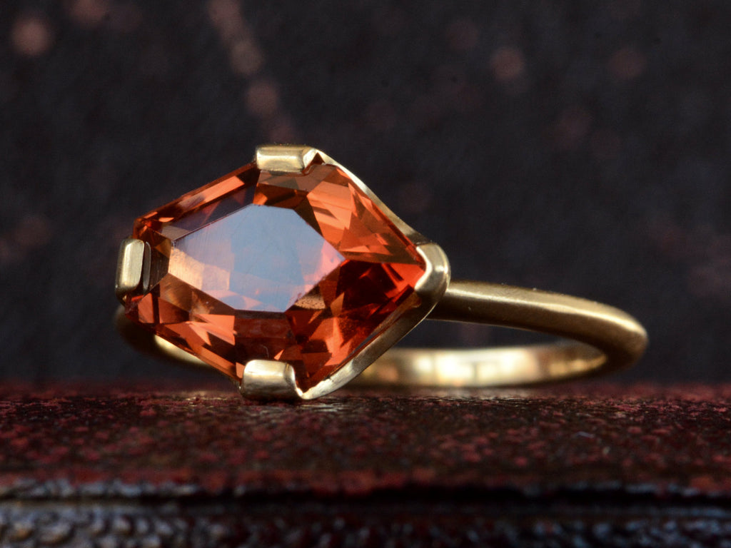 Right, angled view of shield shaped orangey red stone set in 18K yellow gold, on dark background.