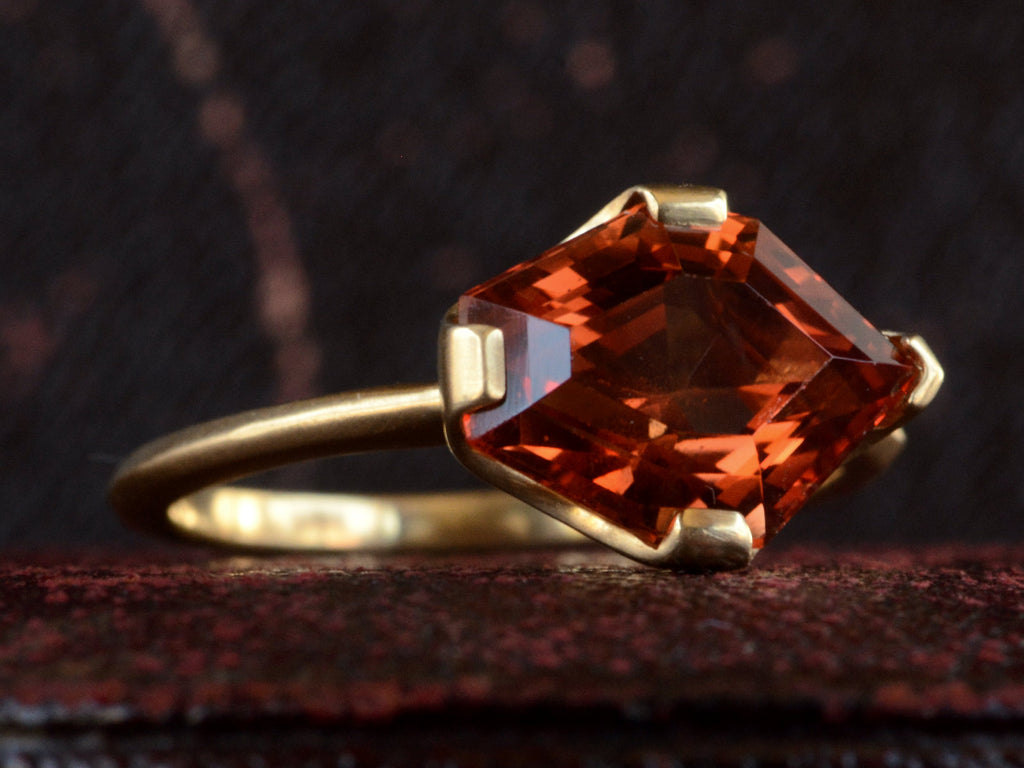 Left, angled view of shield shaped orangey red stone set in 18K yellow gold, on dark background.