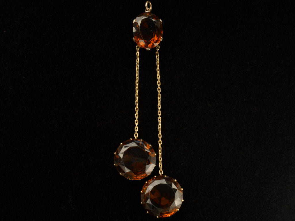 A massive gold Arts & Crafts movement pendant with an oval amber-colored garnet top and two large round amber-colored garnet drops. On black background.