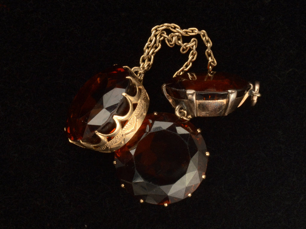 Detail of settings of a massive gold Arts & Crafts movement pendant with an oval amber-colored garnet top and two large round amber-colored garnet drops. On black background.