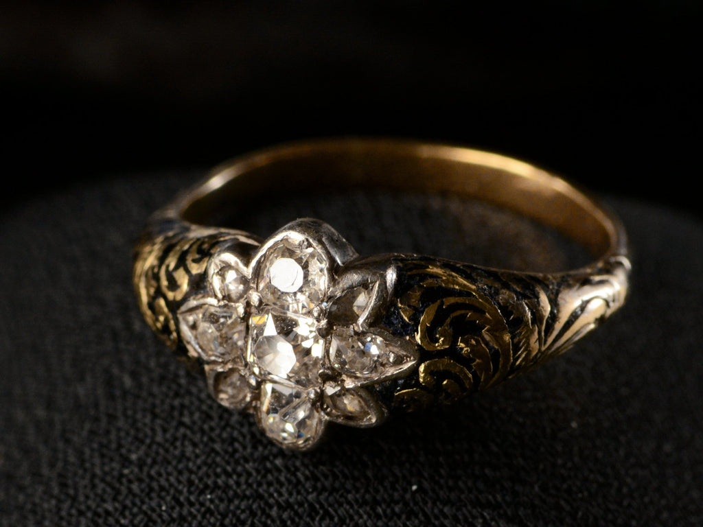 Elevated right angled view of antique c1840 diamond cluster ring in silver topped yellow gold with black enamel scroll designs on the shoulders, shown on dark background.