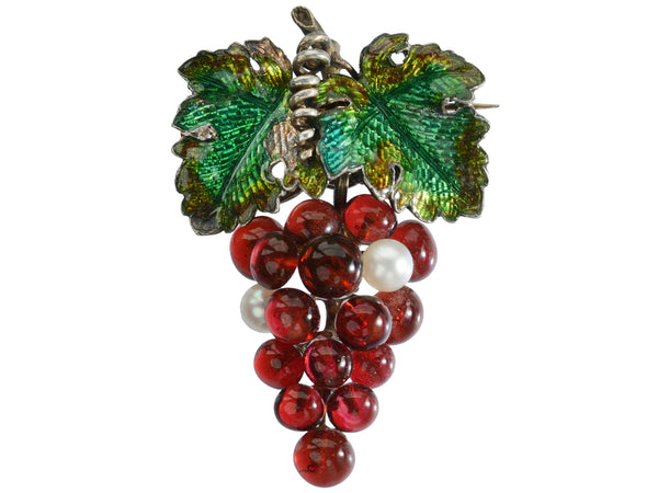 An antique French brooch with enameled green grape leaves and a cluster of red glass and pearl grapes. Shown on white background.