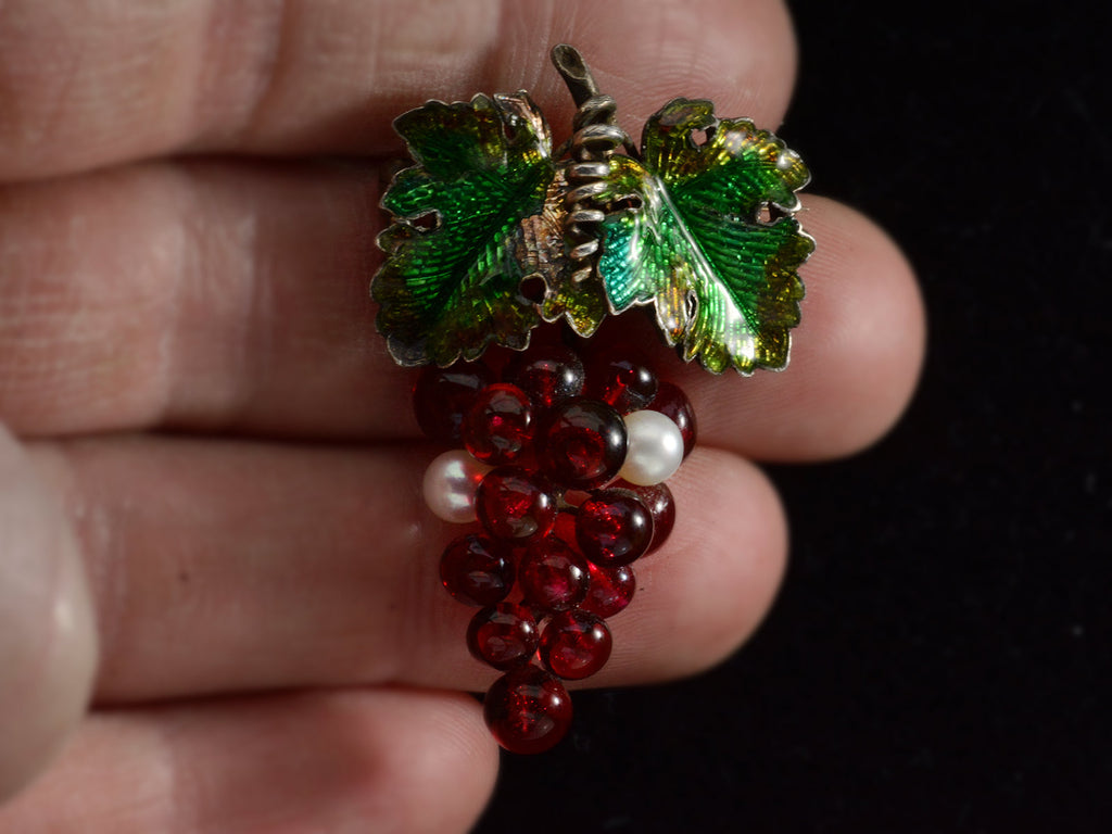 An antique French brooch with enameled green grape leaves and a cluster of red glass and pearl grapes. Shown on hand.