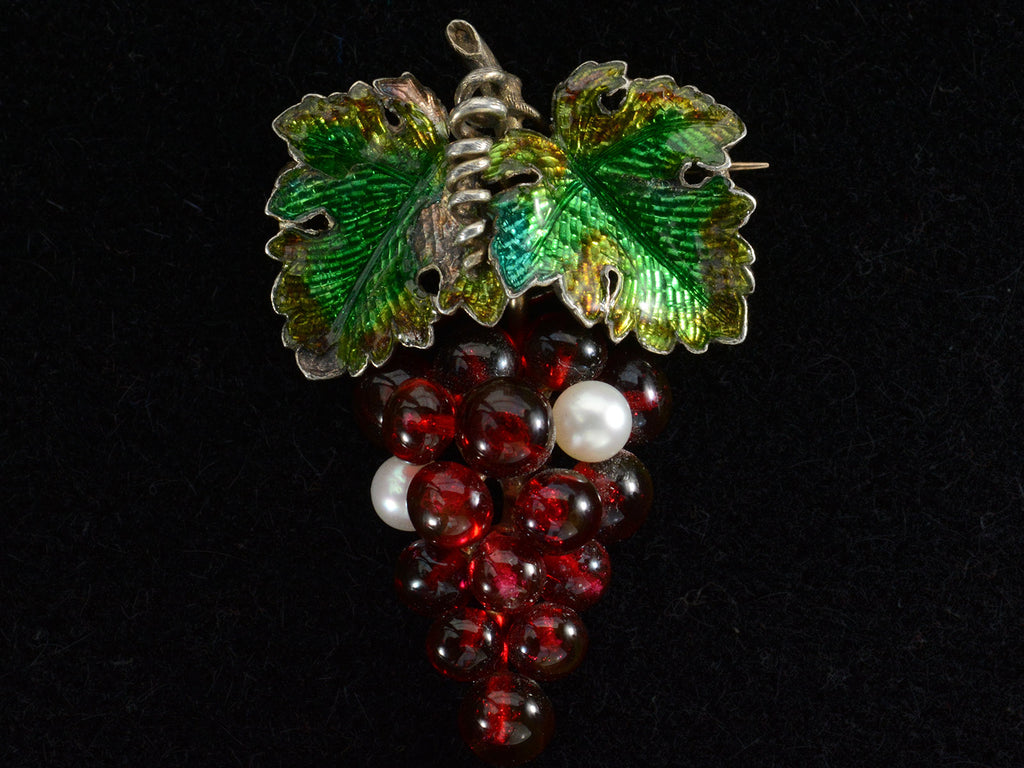 An antique French brooch with enameled green grape leaves and a cluster of red glass and pearl grapes. Shown on black background.