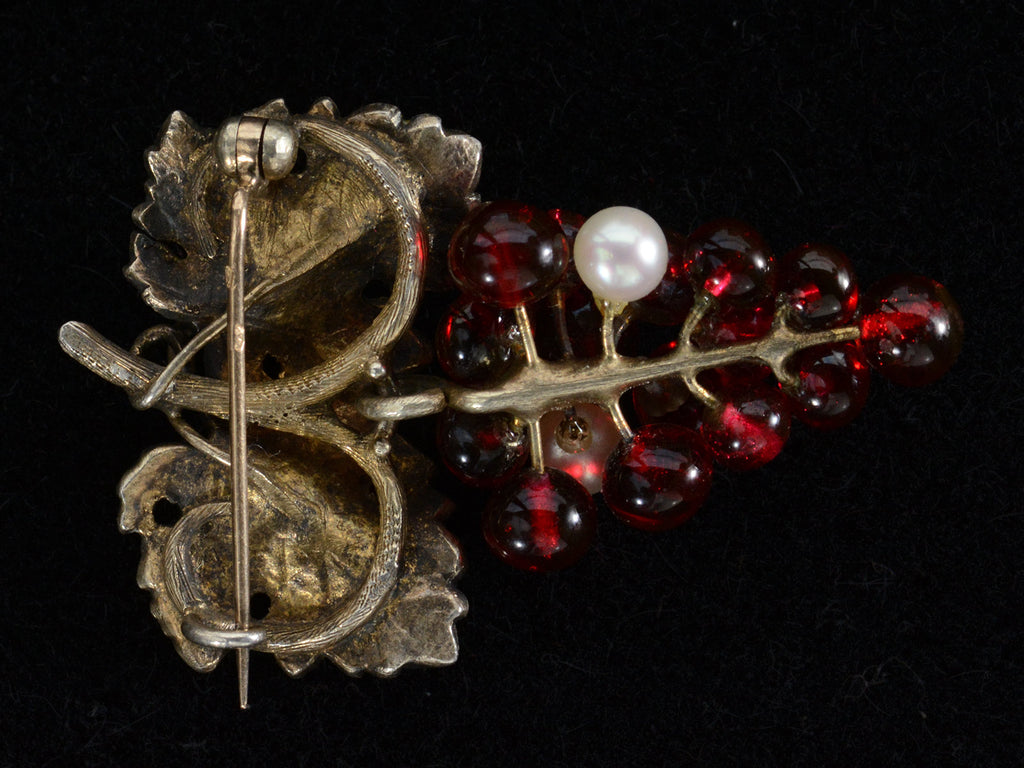 Reverse side of antique French brooch with enameled green grape leaves and a cluster of red glass and pearl grapes. Shown on black background.