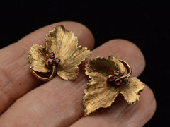 thumbnail of Pair of grape leaf shaped earrings in 18K yellow gold with ruby centers, shown on hand for scale.