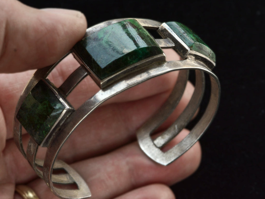 A hand holding a wide silver cuff bracelet with four rectangular green malachite inlays, shown against a dark background.