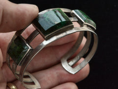 thumbnail of A hand holding a wide silver cuff bracelet with four rectangular green malachite inlays, shown against a dark background.