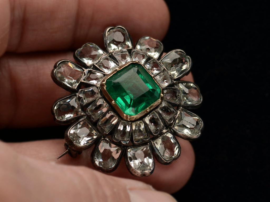 Hand holding a 19th century silver and gold brooch with a green crystal surrounded by petals of clear white faceted crystals