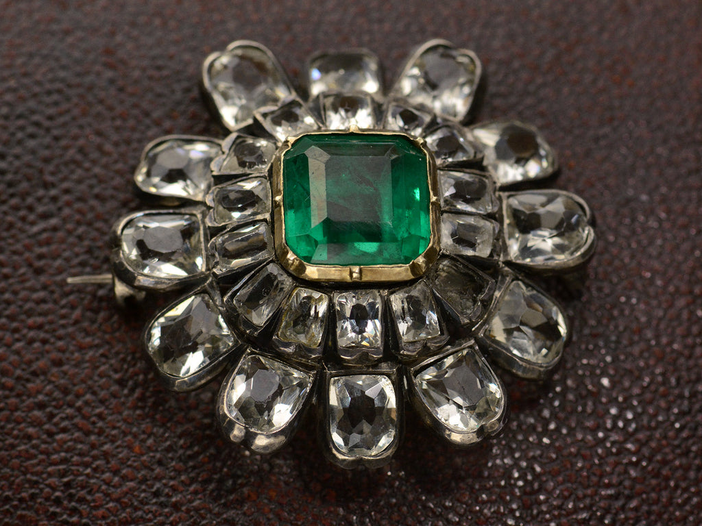 Angled view of 19th century silver and gold brooch with a green crystal surrounded by petals of clear white faceted crystals, shown on dark textured background.