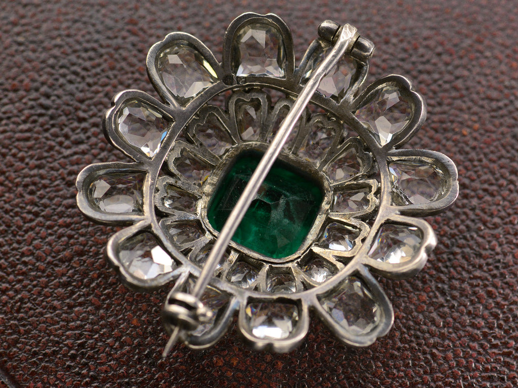 Reverse side view of 19th century silver and gold brooch with a green crystal surrounded by petals of clear white faceted crystals, shown on dark textured background.