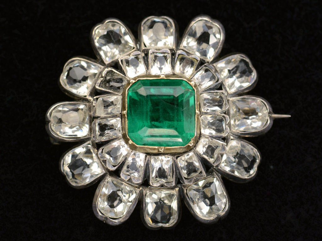 Face on view of 19th century silver and gold brooch with a green crystal surrounded by petals of clear white faceted crystals, shown on dark background.