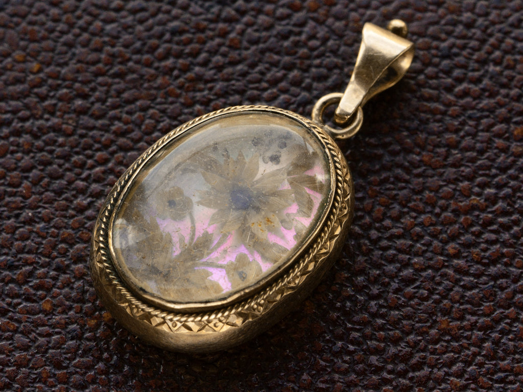 Angled view of Victorian gold mourning pendant with blond hair arrange in a floral design on iridescent mother of pearl, shown on dark textured background.