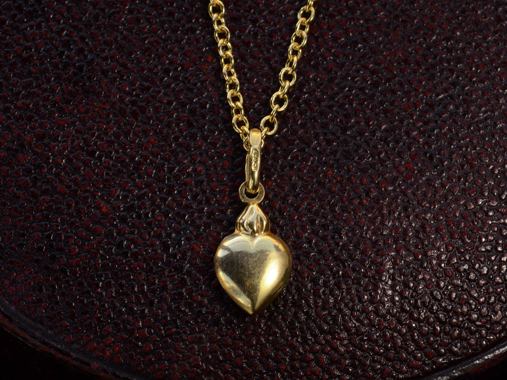 Yellow gold flaming heart shaped charm  hanging on a cable chain, shown on dark textured background.  A stamp on the bale reads "750."