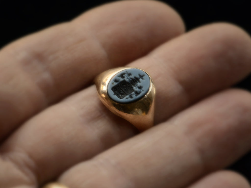 An antique yellow gold signet ring with an oval black and blue chalcedony intaglio with an armorial coat of arms (a cross on top of a crown on top of a shield with an axe and a fox), shown on finger.