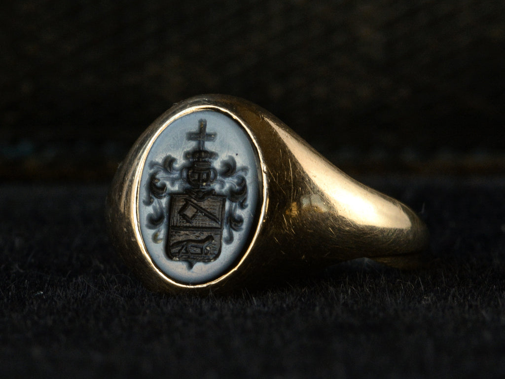 Angled view of antique yellow gold signet ring with an oval black and blue chalcedony intaglio with an armorial coat of arms (a cross on top of a crown on top of a shield with an axe and a fox), shown on dark background.