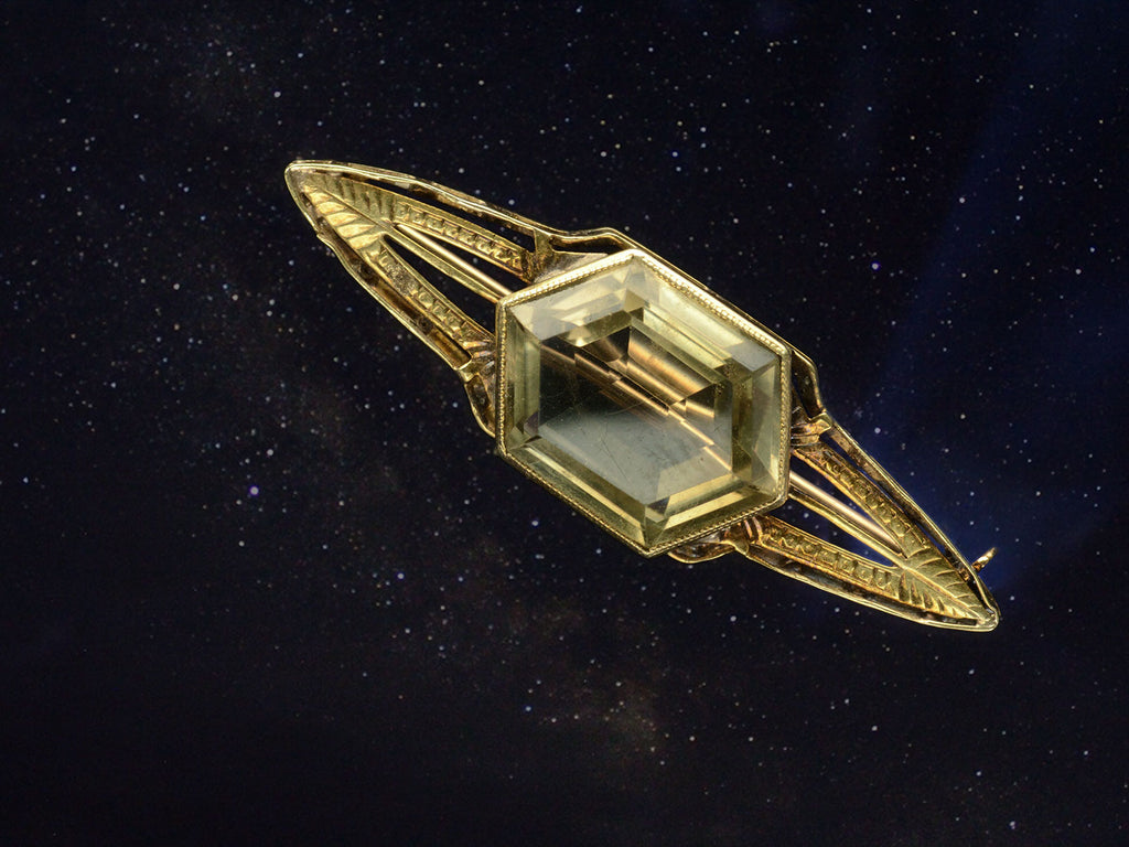 Antique Art Deco brooch with hexagonal citrine and gold wings, resembling a flying saucer, shown superimposed on starry night sky.