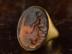 thumbnail of Right angled view of antique yellow gold signet ring with amber glass intaglio of classical scene (Eros fishing), shown on dark background.