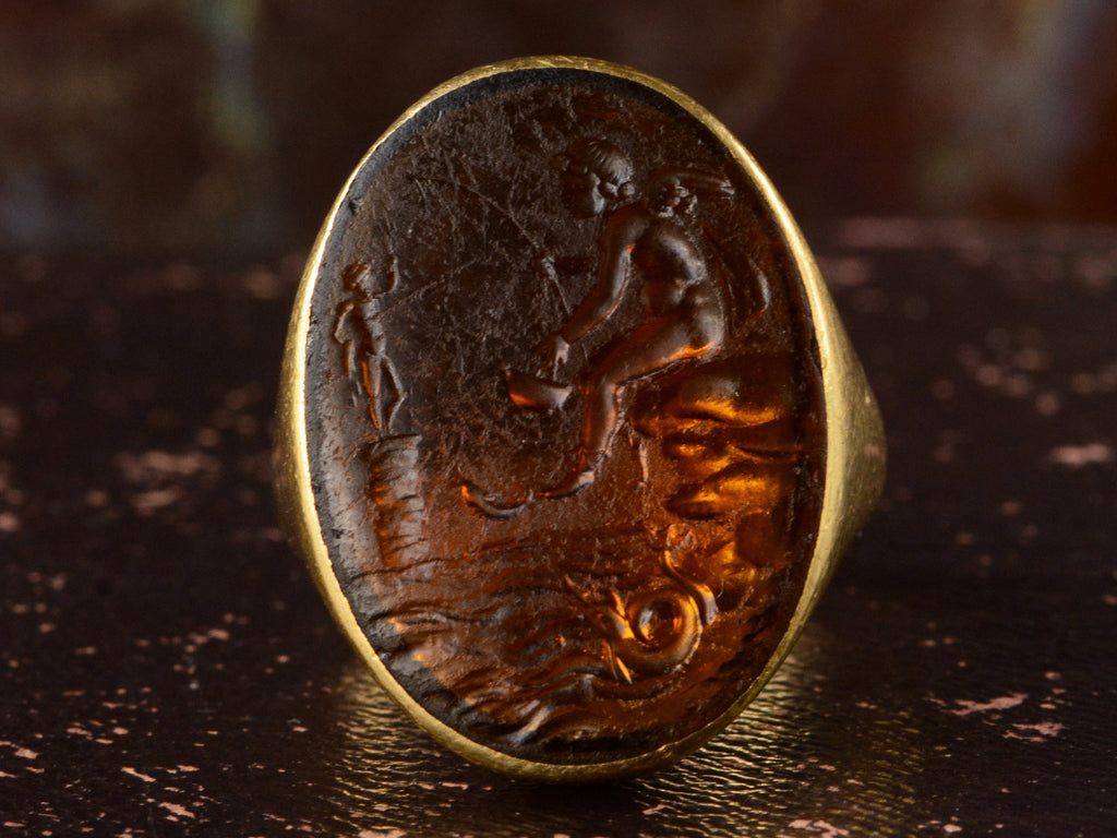 Face profile view of antique yellow gold signet ring with amber glass intaglio of classical scene (Eros fishing), shown on dark background.