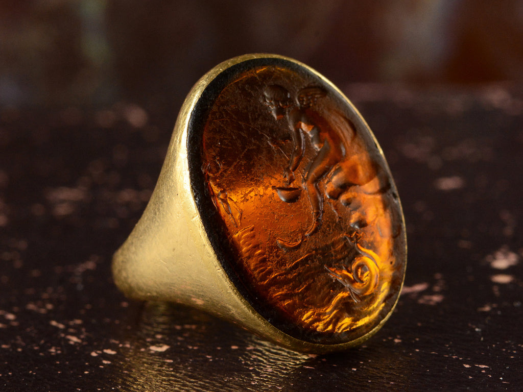 Left angle view of antique yellow gold signet ring with amber glass intaglio of classical scene (Eros fishing), shown on dark background.