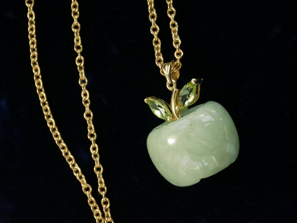 Detail view of light green apple shaped pendant carved from jade with a gold stem and peridot leaves, shown with cable chain on black background.