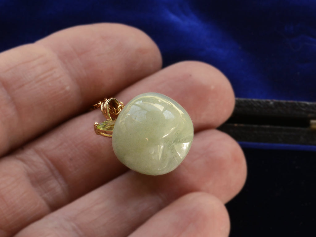 Bottom view of light green apple shaped pendant carved from jade with a gold stem and peridot leaves, shown on hand.