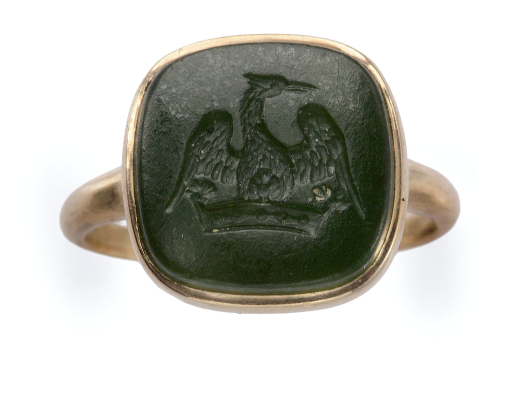 An antique signet ring with a dark green intaglio carved with the image of a crane stretching his wings over a crown, mounted in rosy-yellow gold and shown on white background.