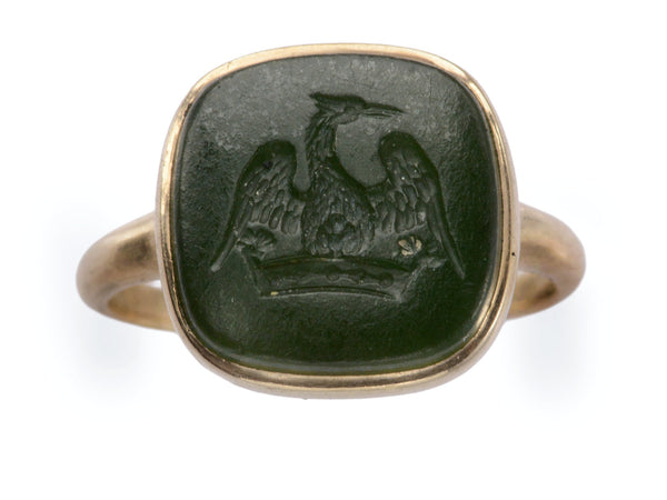 An antique signet ring with a dark green intaglio carved with the image of a crane stretching his wings over a crown, mounted in rosy-yellow gold and shown on white background.