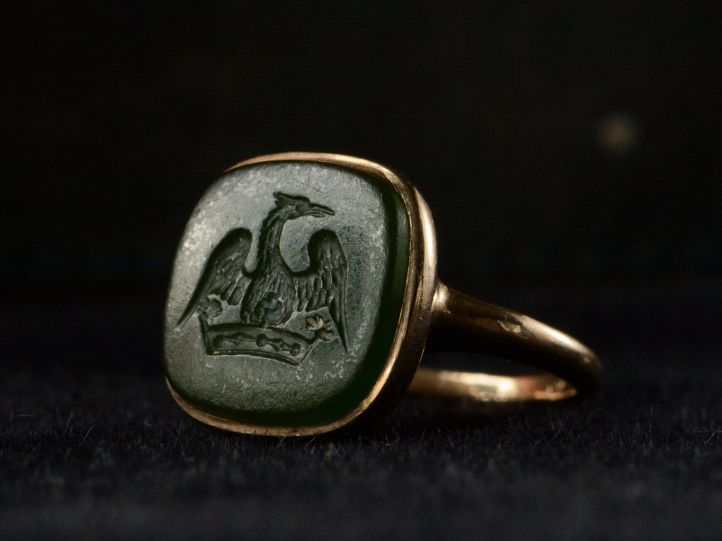 Angled view of antique signet ring with a dark green intaglio carved with the image of a crane stretching his wings over a crown, mounted in rosy-yellow gold and shown on black background.