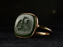 thumbnail of Angled view of antique signet ring with a dark green intaglio carved with the image of a crane stretching his wings over a crown, mounted in rosy-yellow gold and shown on black background.