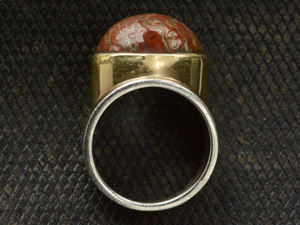 Side profile view of fossilized red jasper cabochon set in gold bezel on silver shank, shown on dark textured background.