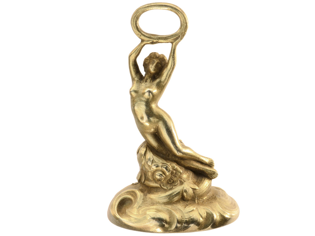 Antique gold watch fob with a nude women resting on a base of vines, flowers and flowing water.  Shown on white background.